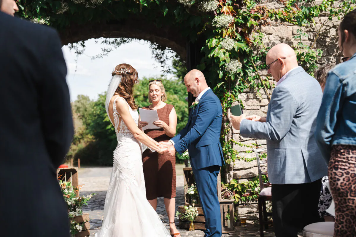 A bride and groom hold hands during an outdoor celebrant-led ceremony in front of a stone archway draped in ivy.