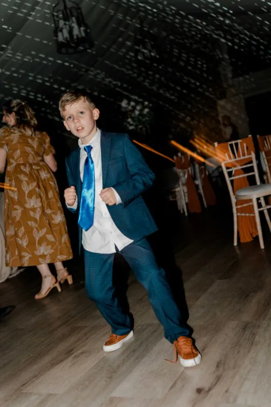 A young page boy in a blue checked suit and vibrant blue tie enthusiastically dances on the wooden floor during the evening wedding reception.
