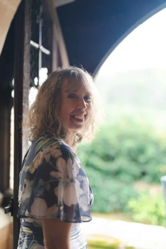 A smiling guest wearing a floral print dress glances back over her shoulder while standing in an arched doorway bathed in soft natural light.