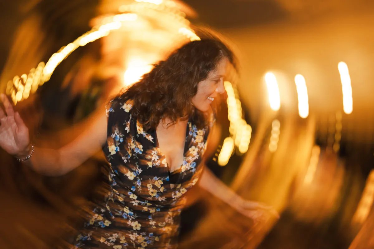A wedding guest with dark curly hair dances energetically under glowing fairy lights, captured with creative motion blur during the evening reception.