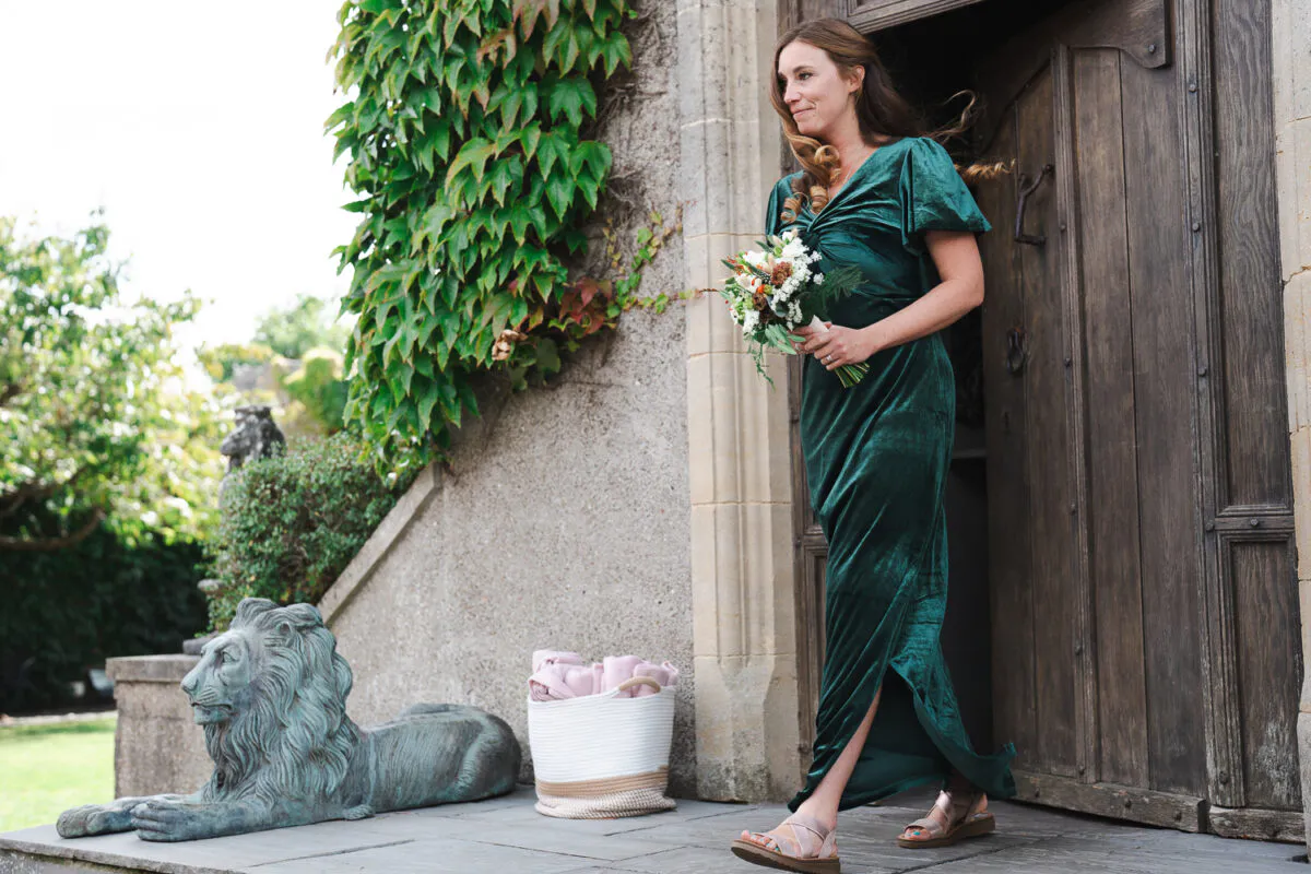 A bridesmaid wearing an emerald green velvet dress and holding a posy bouquet steps out of a stone doorway beside an ivy-covered wall.
