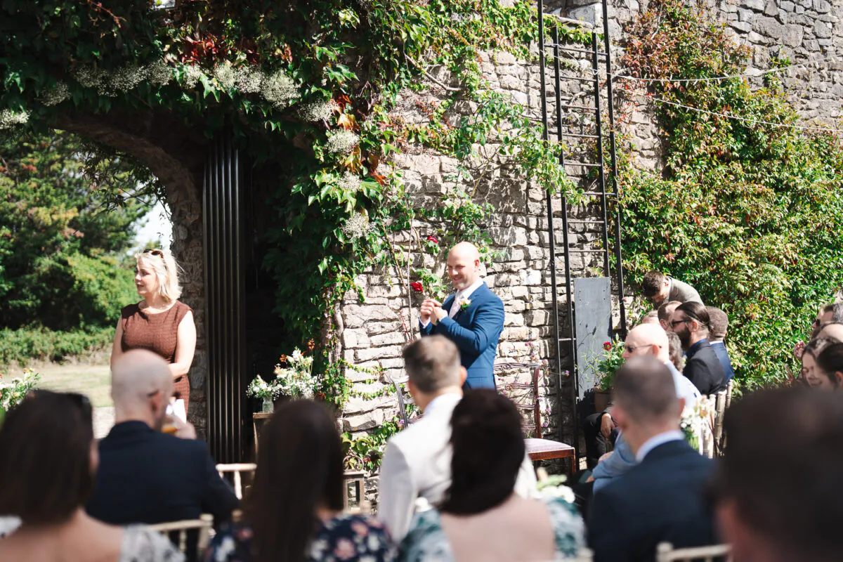 A groom in a blue suit stands waiting at the altar beneath a rustic stone archway covered in lush green ivy during an outdoor wedding ceremony.