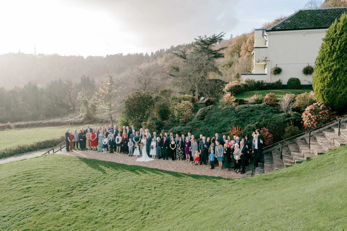 A large wedding party and guests pose for a group portrait on a gravel path outside a country house with scenic rolling hills in the background during golden hour.
