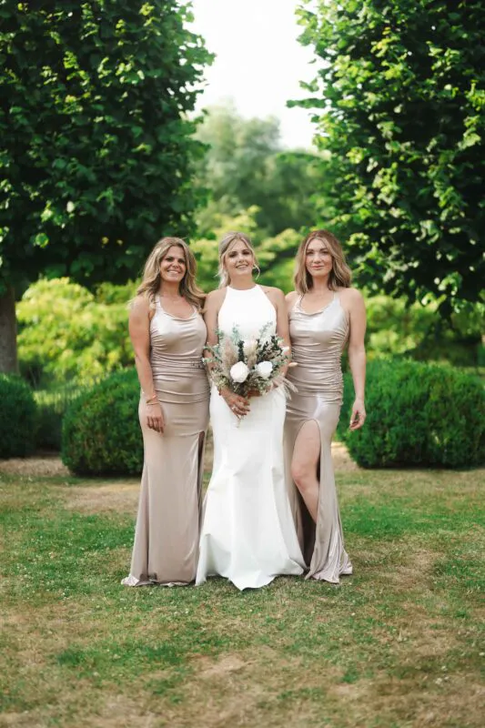 A smiling bride holding a bouquet of white roses and dried grasses poses outdoors alongside two bridesmaids wearing matching metallic gold satin gowns with thigh splits.