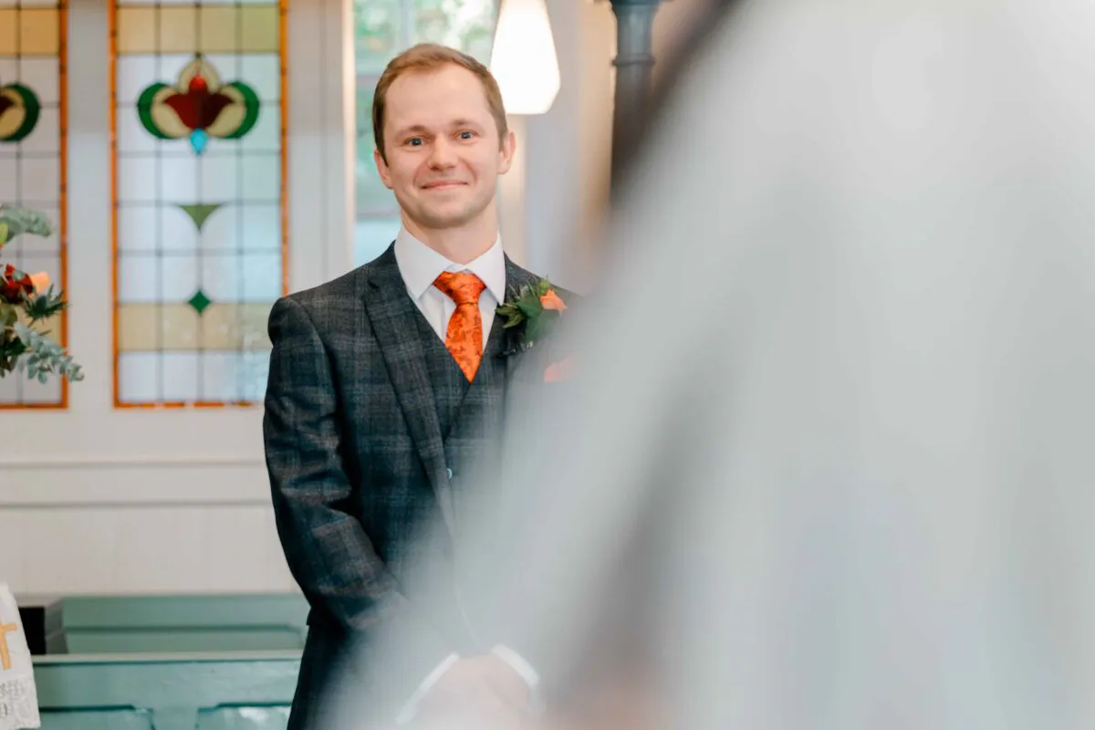 A smiling groom wearing a checked three-piece suit and orange patterned tie waits at the altar during an indoor ceremony.