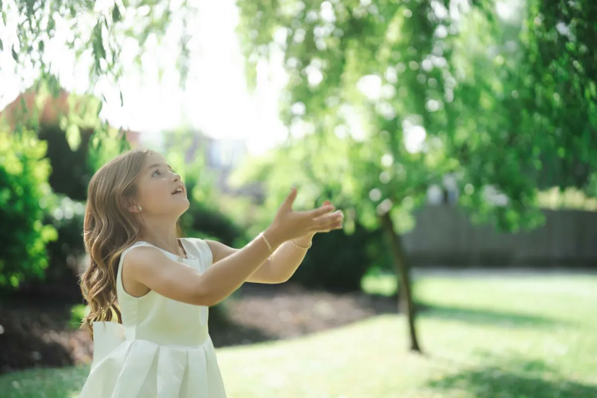 A young flower girl looks up happily with outstretched hands beneath a leafy willow tree on a bright wedding day.