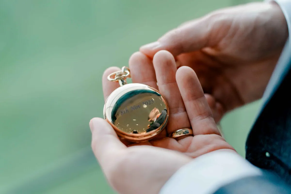 A groom gently holds a polished gold pocket watch engraved with a wedding date in the palm of his hands.