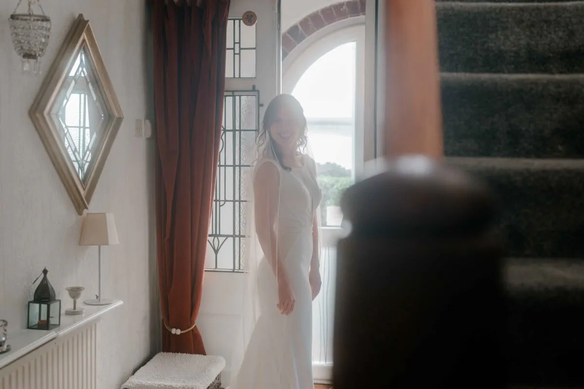 A smiling bride wearing a simple crepe column dress and sheer veil looks back towards the camera while standing in an interior doorway filled with natural light.