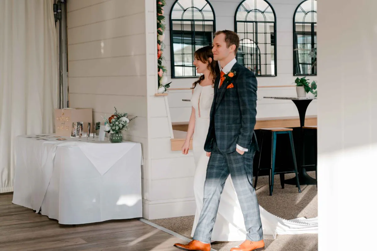 Newlywed bride and groom walk together indoors, with the groom wearing a checked suit and the bride in a sleek crepe gown.