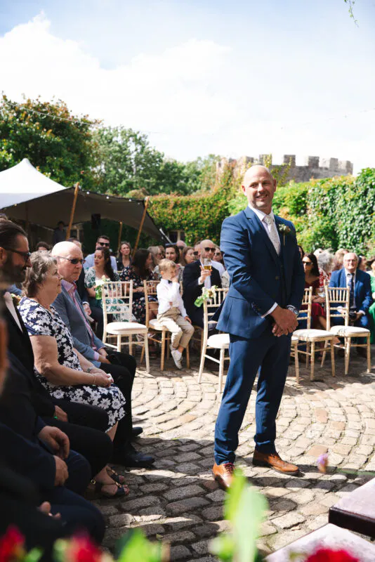 A smiling groom in a blue suit stands waiting at the altar during an outdoor ceremony in a cobbled courtyard.
