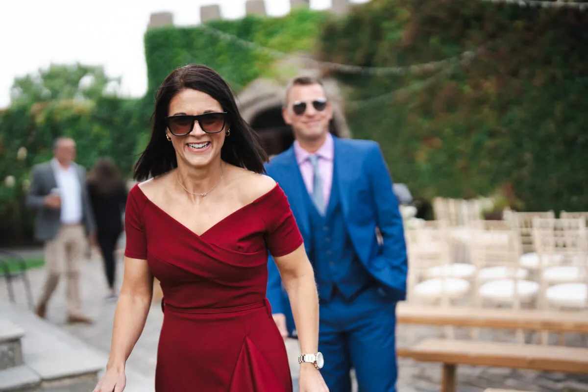 A joyful woman wearing a burgundy dress and sunglasses walks outdoors with a man in a blue suit following closely behind near a ceremony seating area.