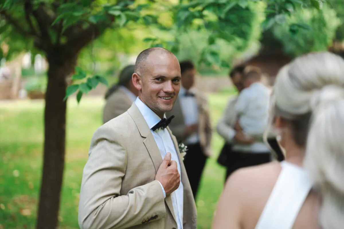 A smiling groom wearing a beige suit and a black bow tie looks back over his shoulder outdoors while guests mingle in the background.