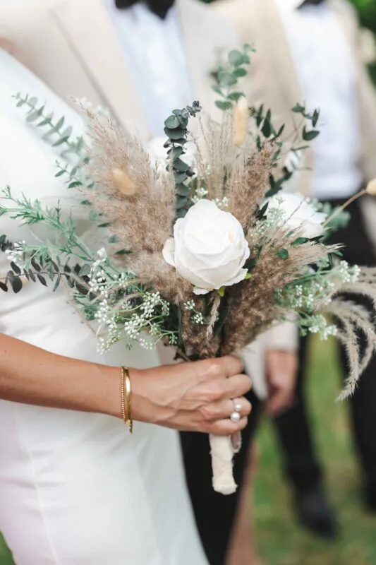 A close-up shot of a bride holding a textured hand-tied bouquet featuring white roses, dried pampas grass, bunny tails, and eucalyptus foliage.