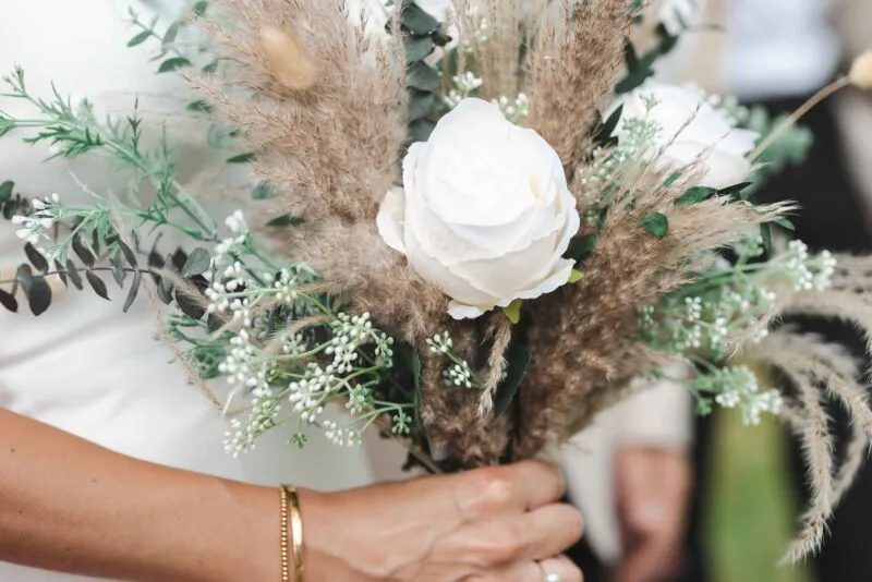 A close-up shot of a bride holding a textured hand-tied bouquet featuring white roses, dried pampas grass, bunny tails, and eucalyptus foliage.