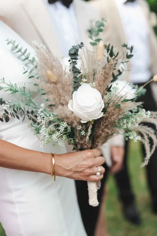 A close-up shot of a bride holding a textured hand-tied bouquet featuring white roses, dried pampas grass, bunny tails, and eucalyptus foliage.