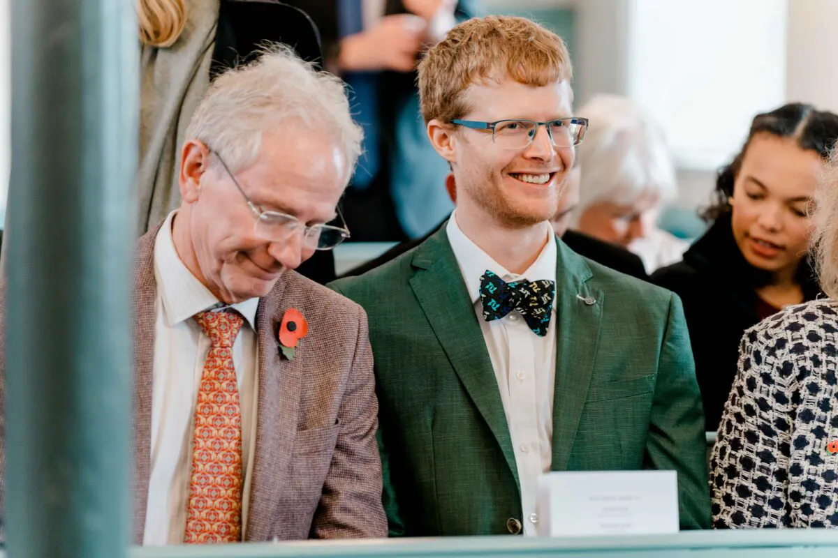A smiling guest wearing a green suit and patterned bow tie sits in a church pew next to an older gentleman during a wedding ceremony.