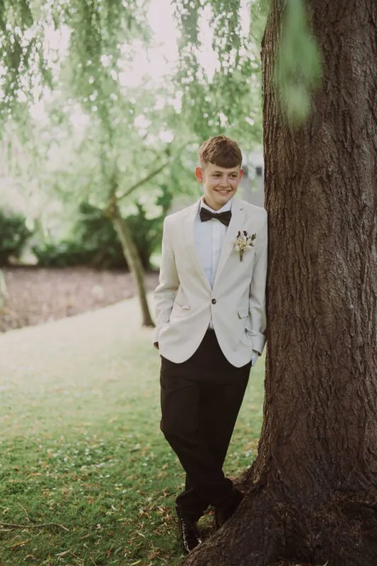 A smiling young page boy wearing a cream suit jacket, black trousers, and a bow tie leans casually against a large tree trunk in a lush woodland setting.