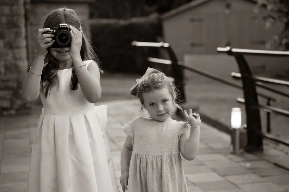 Two young girls in dresses attend an outdoor wedding reception, with one playfully holding up a camera and the other flashing a peace sign.