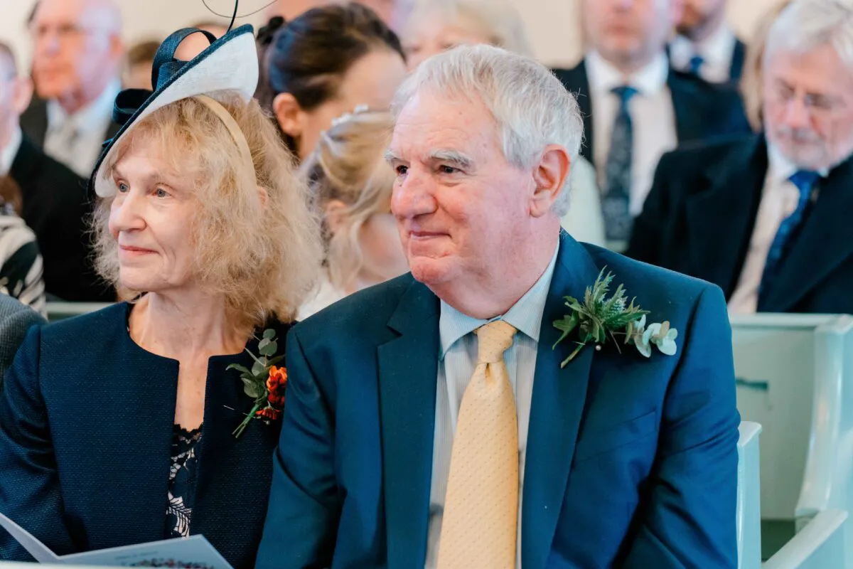 An older couple sitting together in a church pew during a wedding ceremony, with the father of the bride wearing a blue suit and buttonhole.