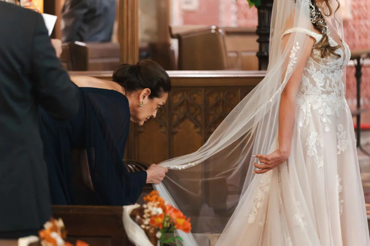 A mother gently arranging the delicate lace-trimmed veil of a bride wearing an off-the-shoulder wedding gown inside a traditional church.