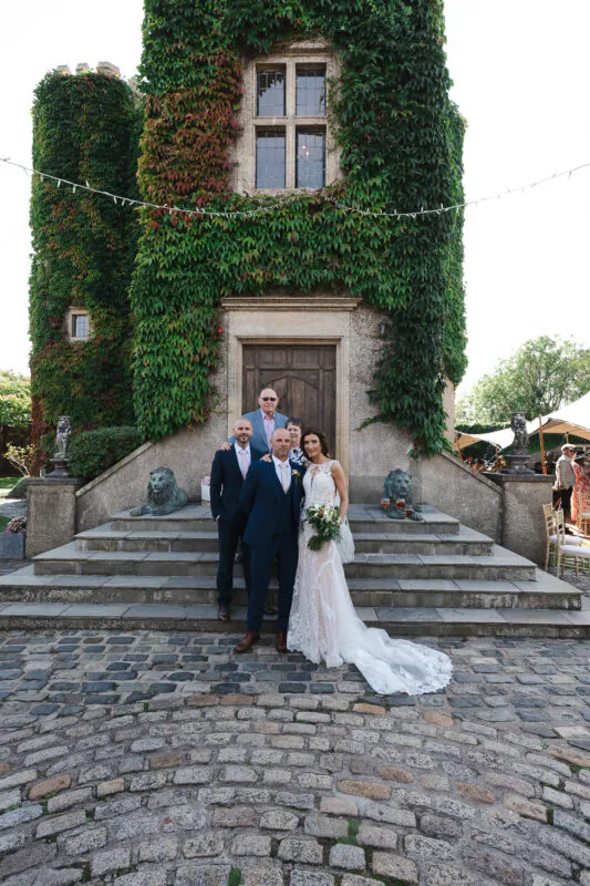 A bridal party and family members pose for a group photo outside a historic ivy-clad stone castle with a cobbled courtyard.