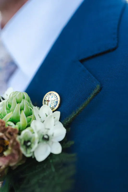 Close-up of a groom's blue suit jacket featuring a floral boutonniere adorned with white blossoms, green foliage, and an Alfa Romeo lapel pin.