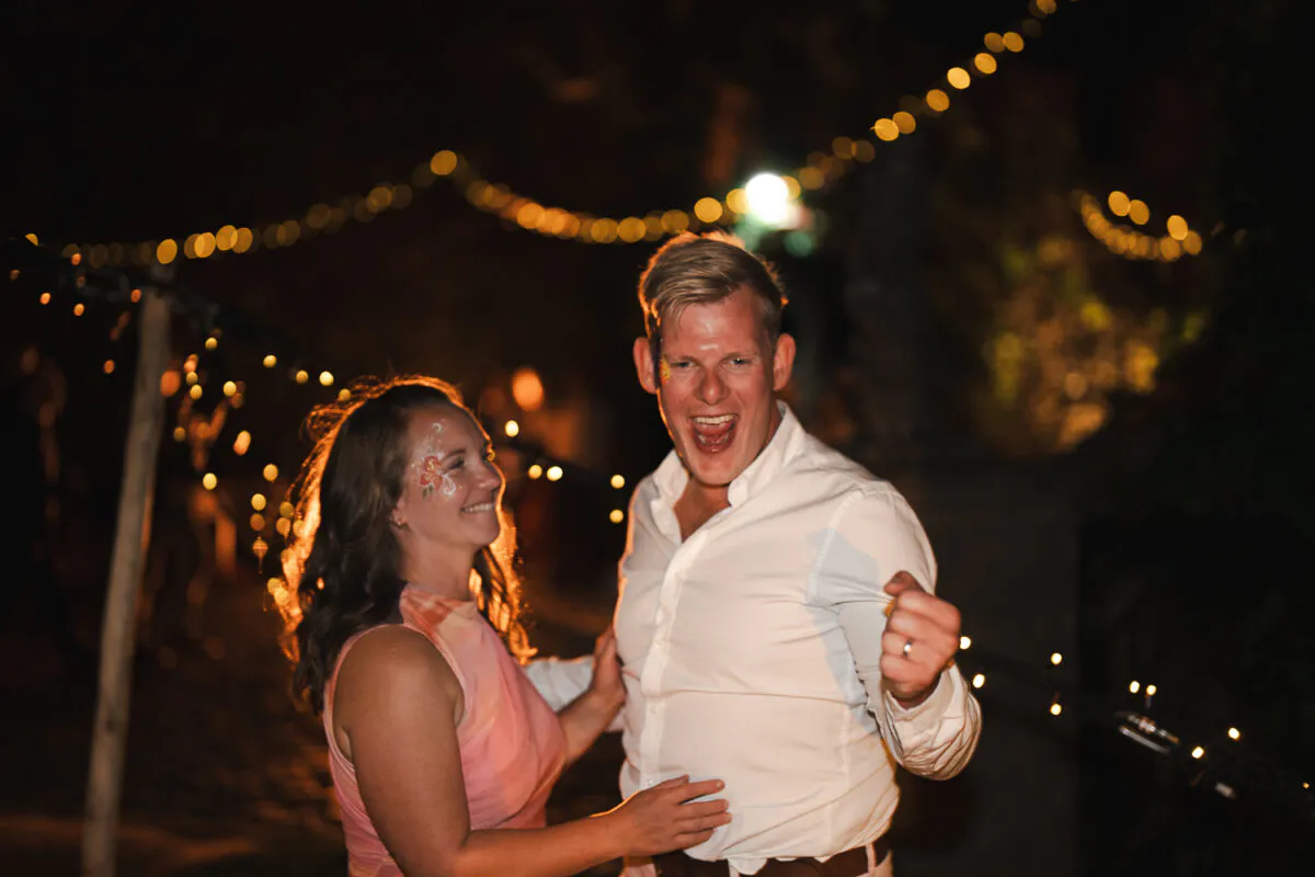 A bride and groom laugh enthusiastically and dance outdoors at night beneath a canopy of twinkling festoon lights.
