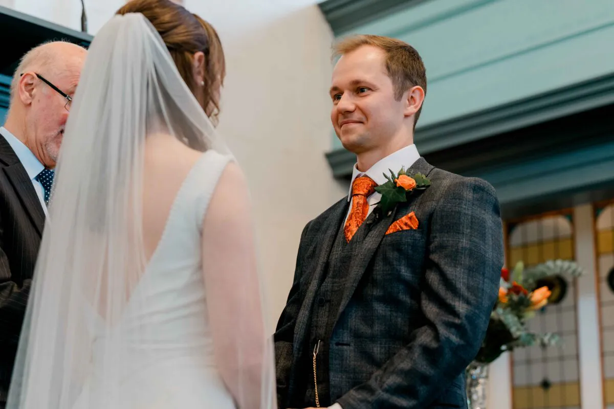 The groom smiles warmly at his bride during the wedding ceremony, wearing a tweed three-piece suit with an orange patterned tie and coordinating pocket square.