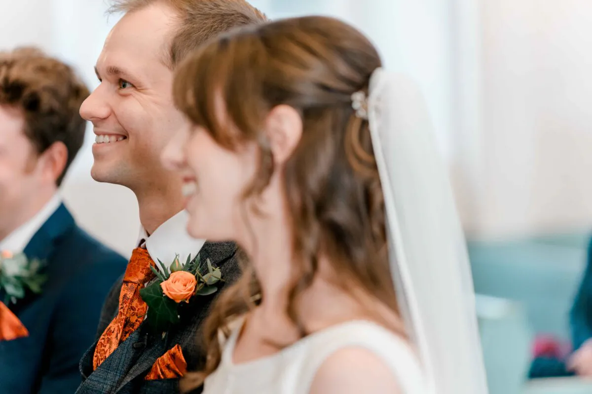 A smiling groom with an orange tie and boutonniere sits next to the bride during the wedding ceremony.