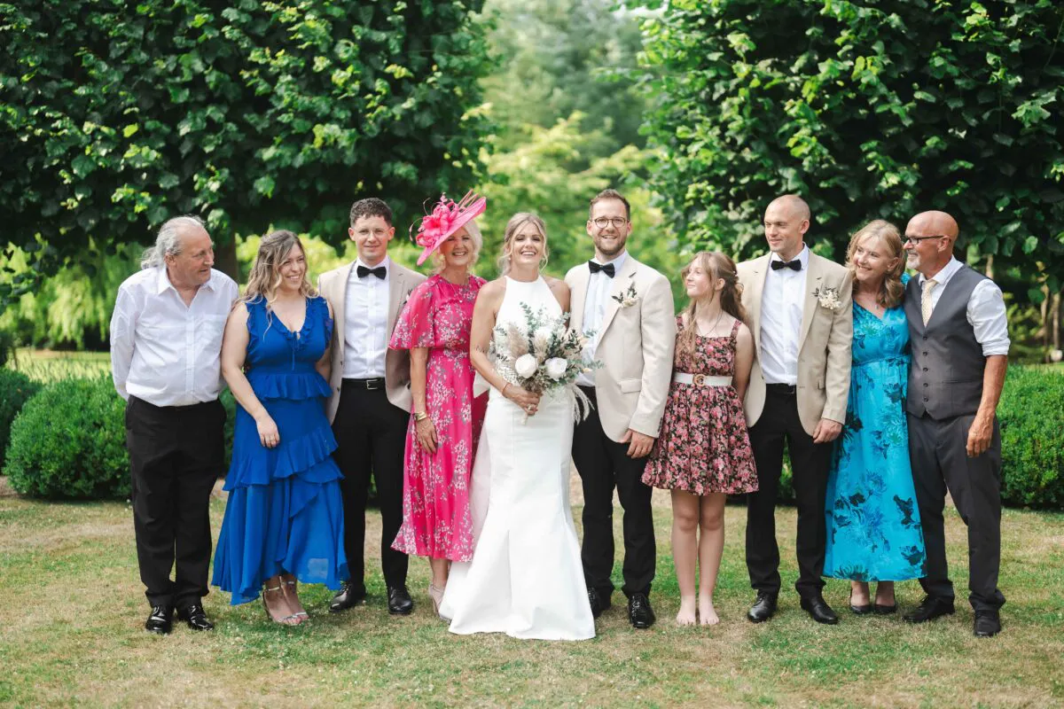A happy wedding group stands outdoors on a lawn against a backdrop of tall green trees, featuring the bride in a white halter neck dress holding a bouquet flanked by family and wedding party members.