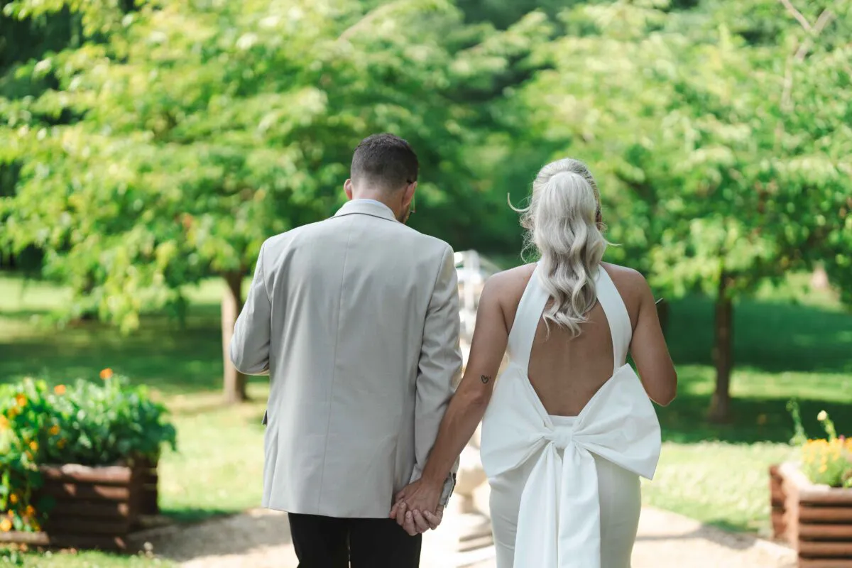 A newlywed couple walking hand-in-hand away from the camera through a lush green garden, featuring the bride in a halterneck gown with an open back and a large white bow detail.