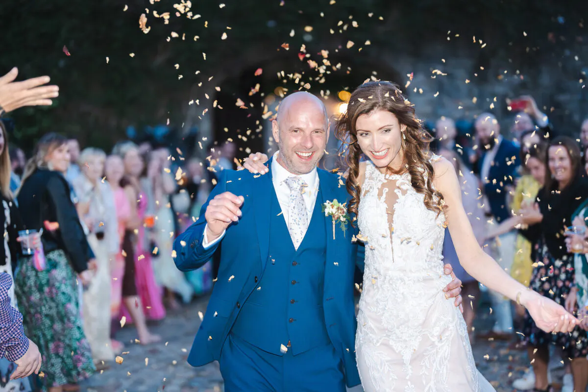 A joyful newlywed couple walks through a shower of confetti amidst cheering guests, with the groom in a sharp blue suit and the bride in an elegant lace gown.