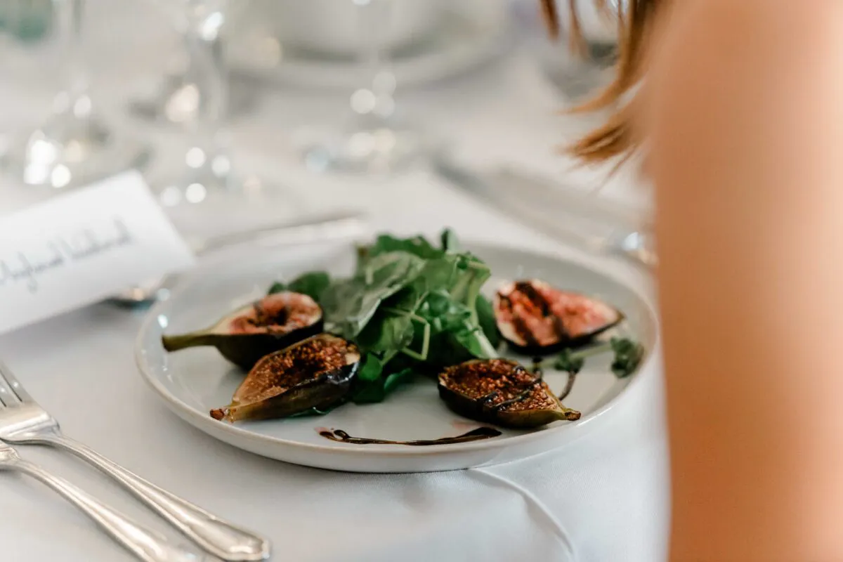 A finely plated wedding starter of fresh figs and salad greens sits on a white table setting with cutlery and a place card.