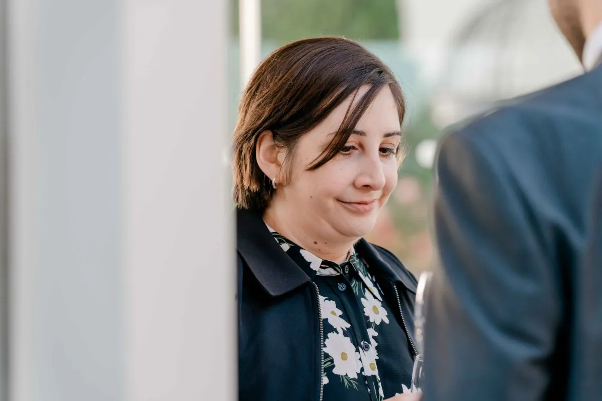 A guest with a short bob hairstyle and dark floral patterned blouse smiles softly while mingling during a wedding reception.