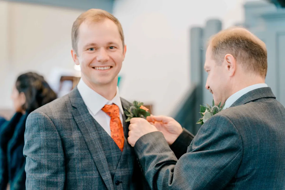 A smiling groom in a grey checked suit and orange patterned tie has his boutonniere adjusted by another man during wedding preparations.