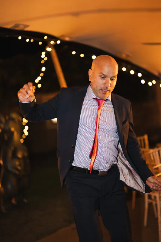 A guest in a navy suit and striped shirt dances energetically at an evening wedding reception under a canopy of twinkling fairy lights.