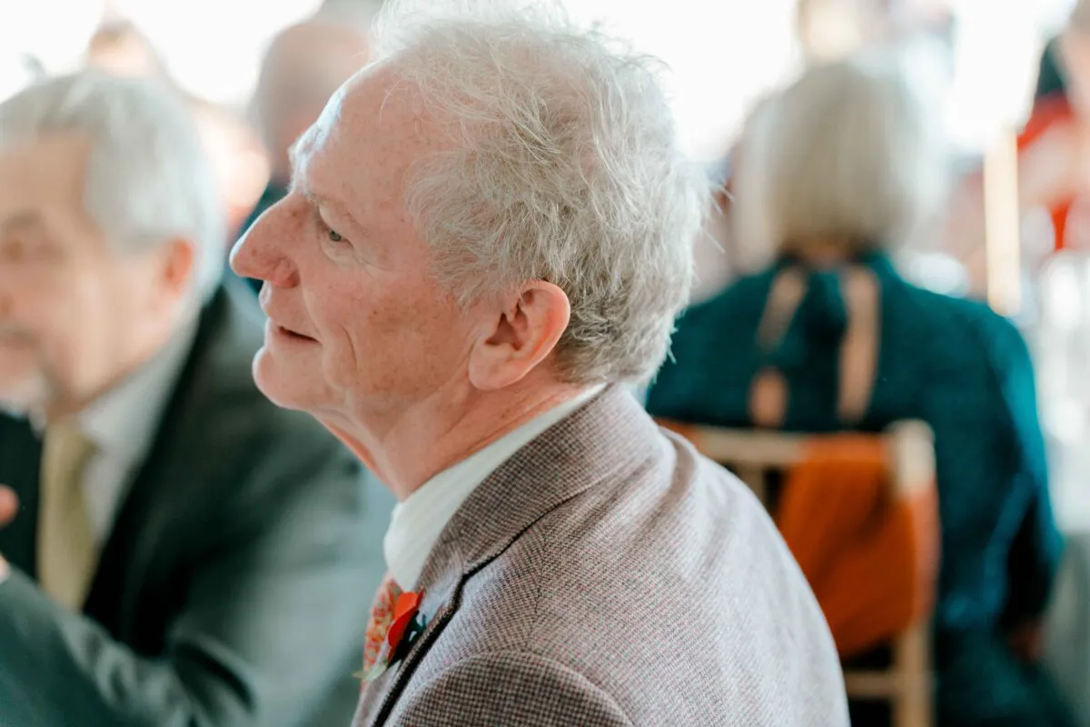 An elderly male guest wearing a tweed suit and a buttonhole laughs candidly during the wedding reception.