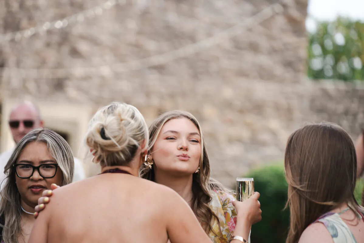 Wedding guests enjoy drinks and laughter outside during the reception against a rustic stone wall backdrop.