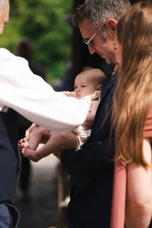 A guest holds a small baby during an outdoor wedding celebration under natural sunlight.