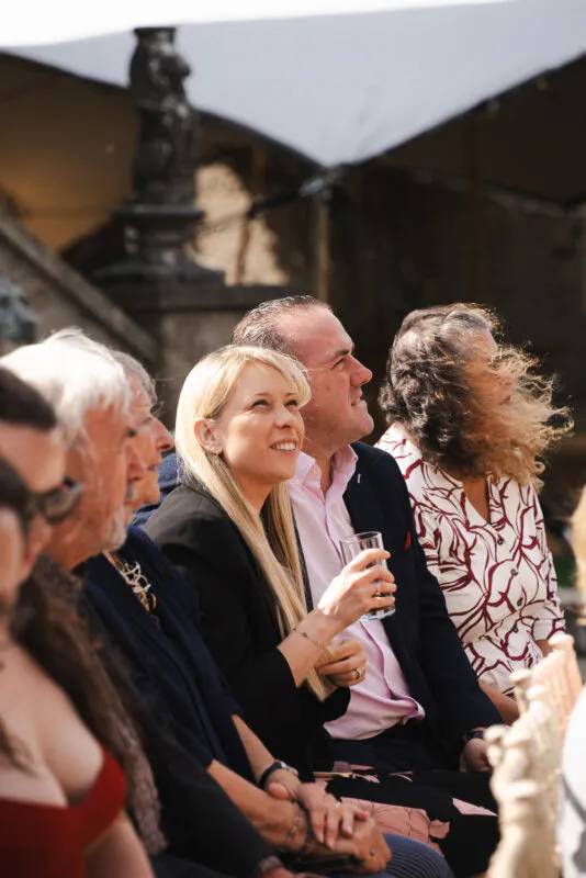 A group of wedding guests seated outdoors in bright sunlight, laughing and enjoying the ceremony together.