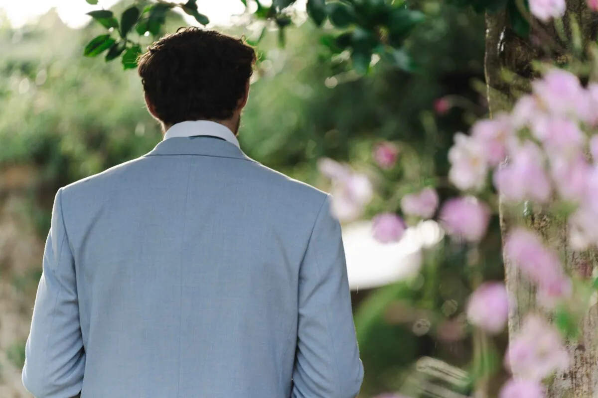 A groom viewed from behind wearing a light grey suit stands outdoors near a tree with pink blossom and lush green foliage in soft natural light.