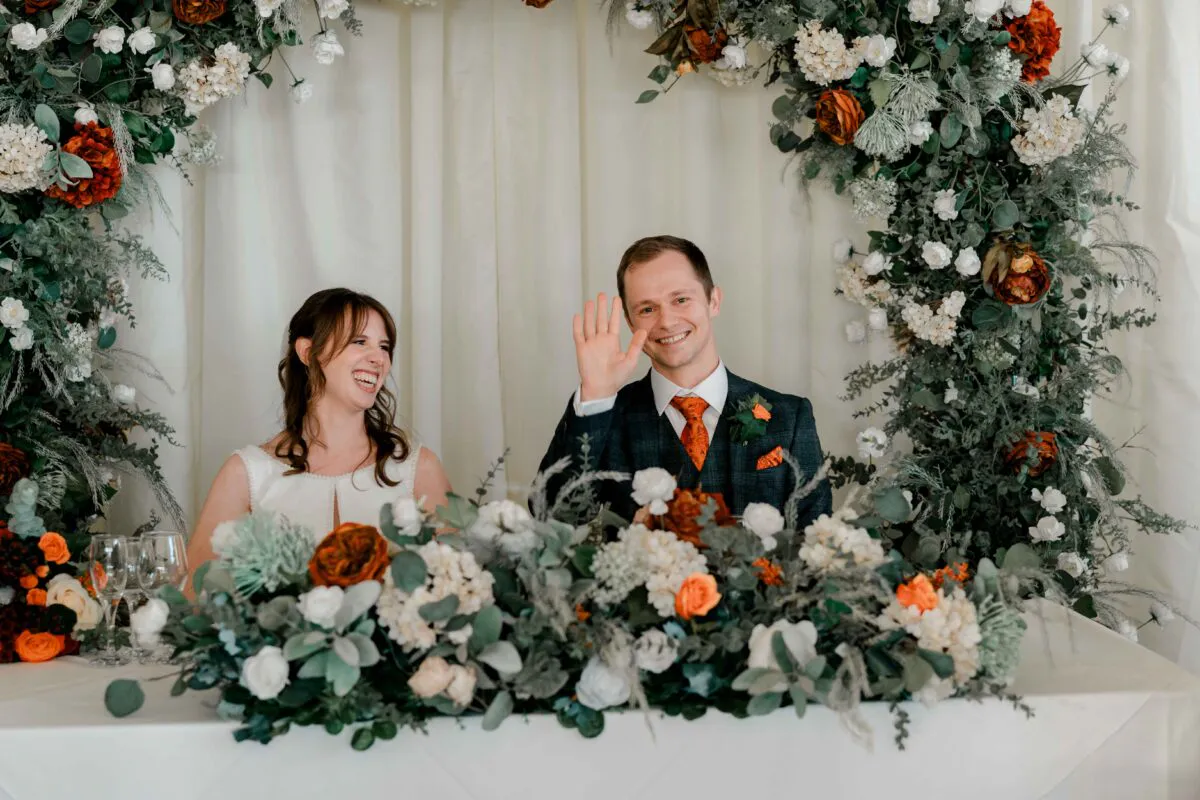 A newlywed couple laughing and waving while seated at the top table, beautifully framed by a large floral arch featuring rich autumn colours.