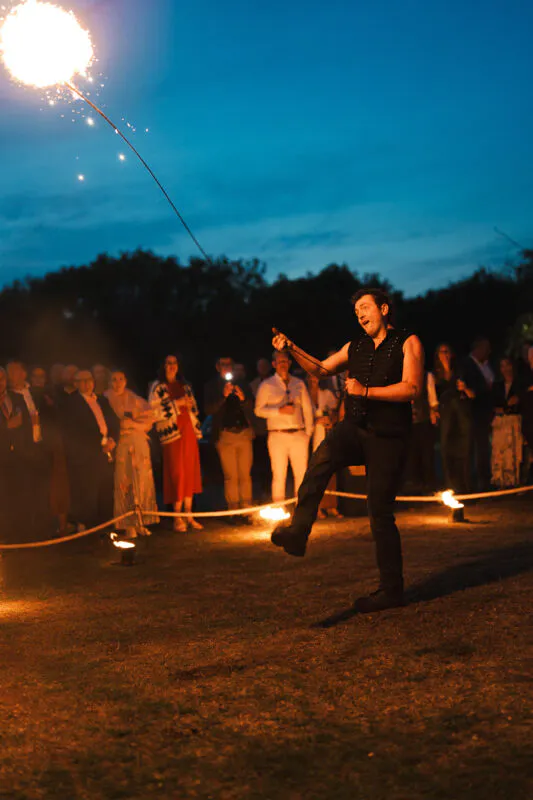 An entertainer swings a lit sparkler outdoors during evening celebrations as wedding guests watch closely under a deep blue twilight sky.