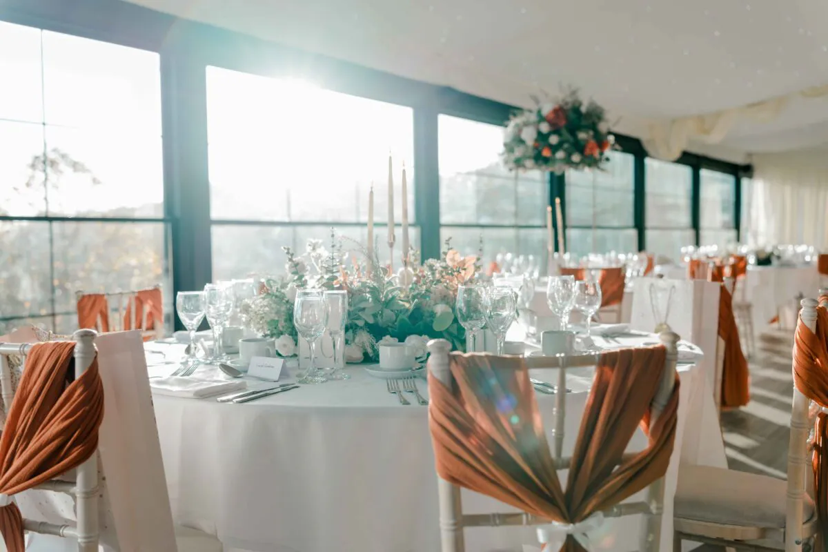 A beautifully styled wedding reception room features round tables adorned with white linens, burnt orange fabric sashes draped over chairs, and elegant floral centrepieces illuminated by natural window light.