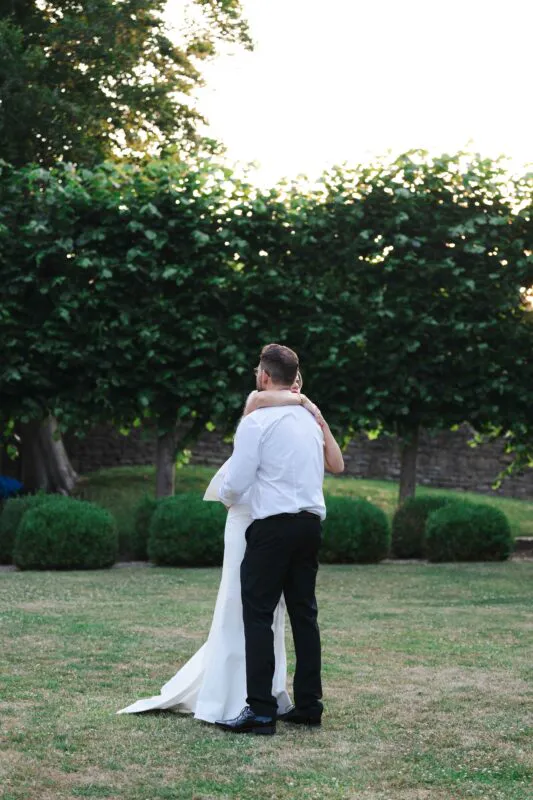 A newlywed couple embraces lovingly on a manicured lawn framed by lush green trees during golden hour.
