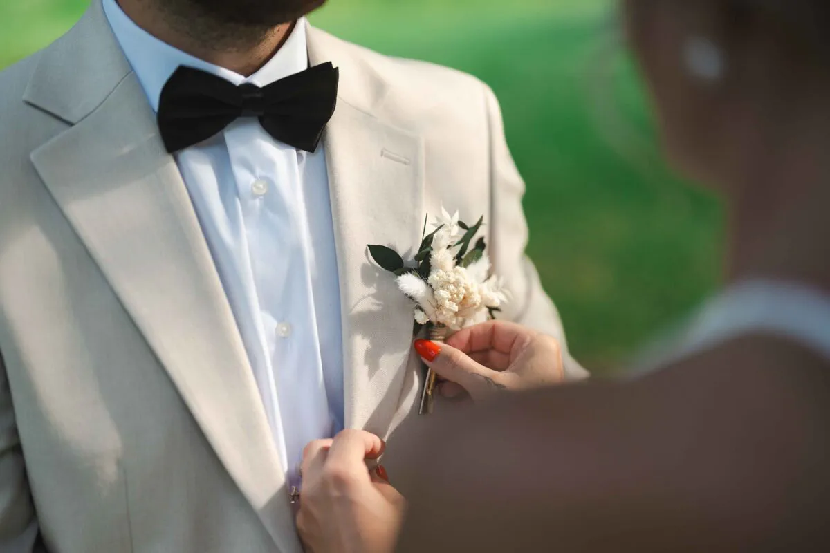A partner carefully pins a dried flower boutonniere to the lapel of a groom wearing a beige suit and a black bow tie.