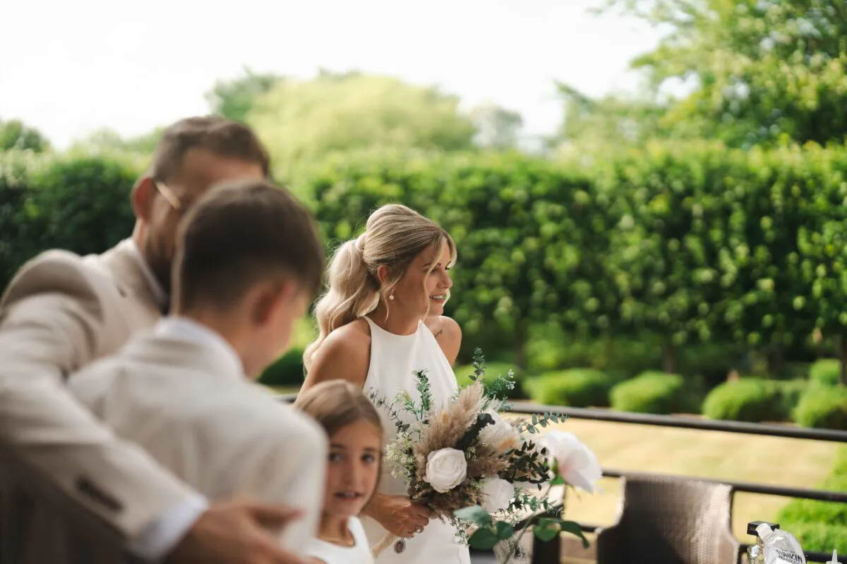 A smiling bride holding a textured bouquet stands outdoors with family members during a sunlit wedding celebration.