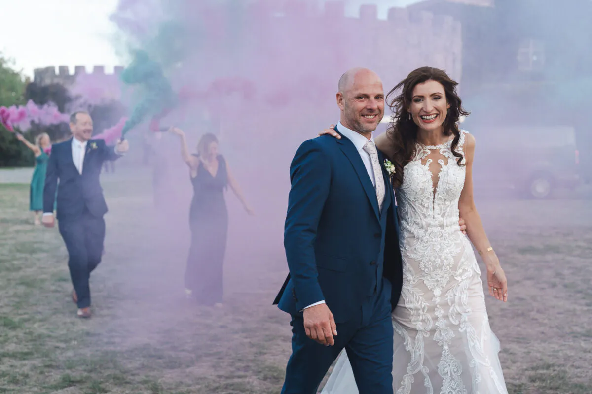A newlywed couple walks joyfully together outside a castle while guests in the background set off vibrant pink and green smoke grenades.