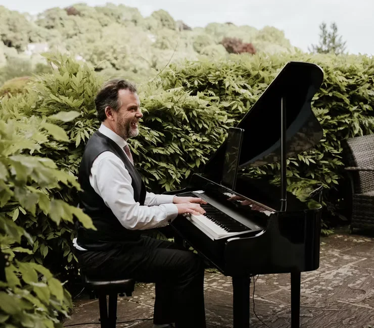 A smiling pianist in a waistcoat and white shirt plays a black grand piano outdoors against a lush green garden backdrop.