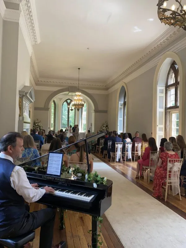 A pianist plays a grand piano in the foreground while wedding guests watch a ceremony unfold at the far end of an elegant, sunlit ballroom.